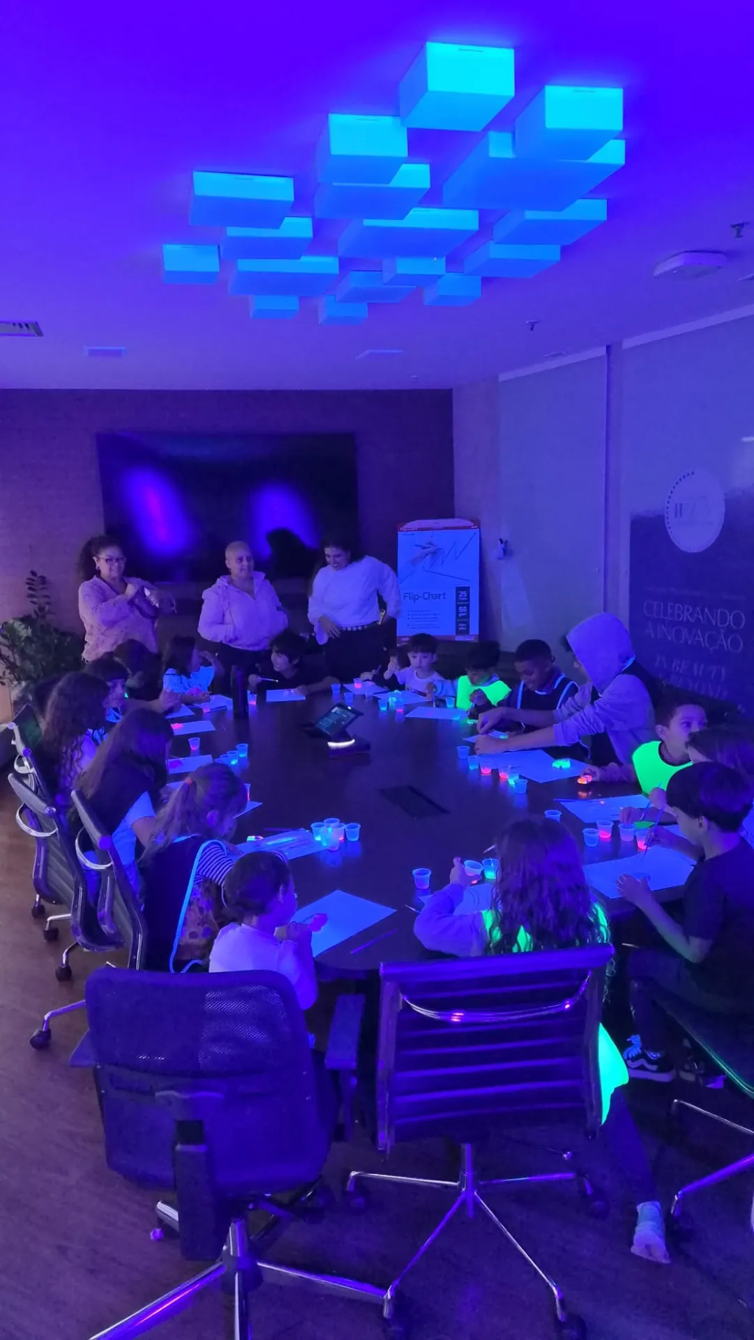 Crianças pintando ao redor de uma mesa de reunião com iluminação neon durante oficina criativa em empresa