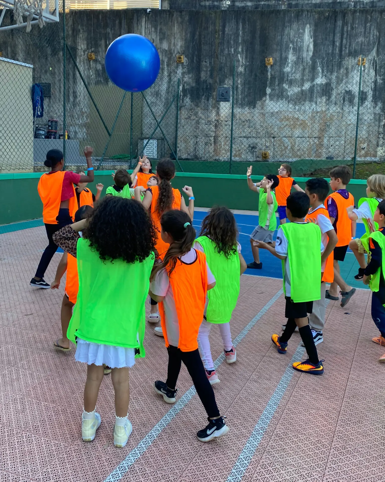 Crianças com coletes laranja e verde disputando bola gigante na quadra do condomínio
