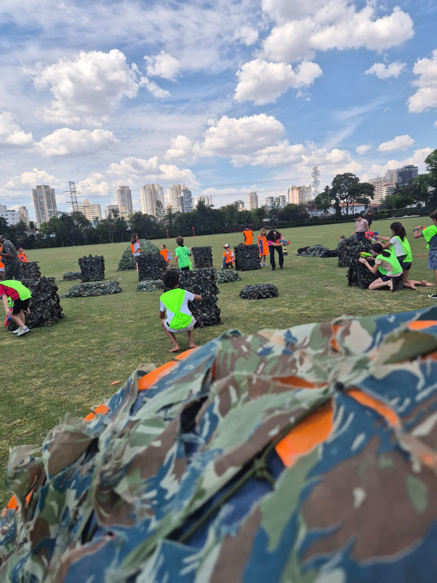 Arena Nerf montada em campo amplo com obstáculos, crianças e recreadores em atividade