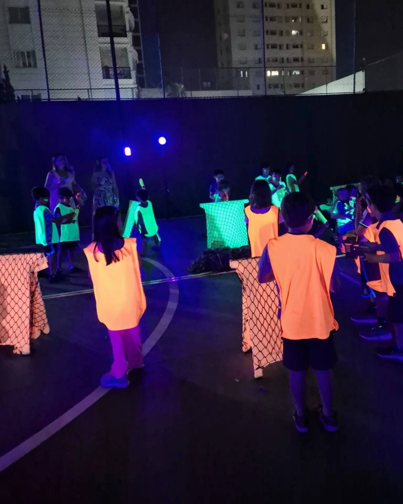 Festa com tema Nerf à noite, com crianças de coletes neon posicionadas atrás das barreiras da arena