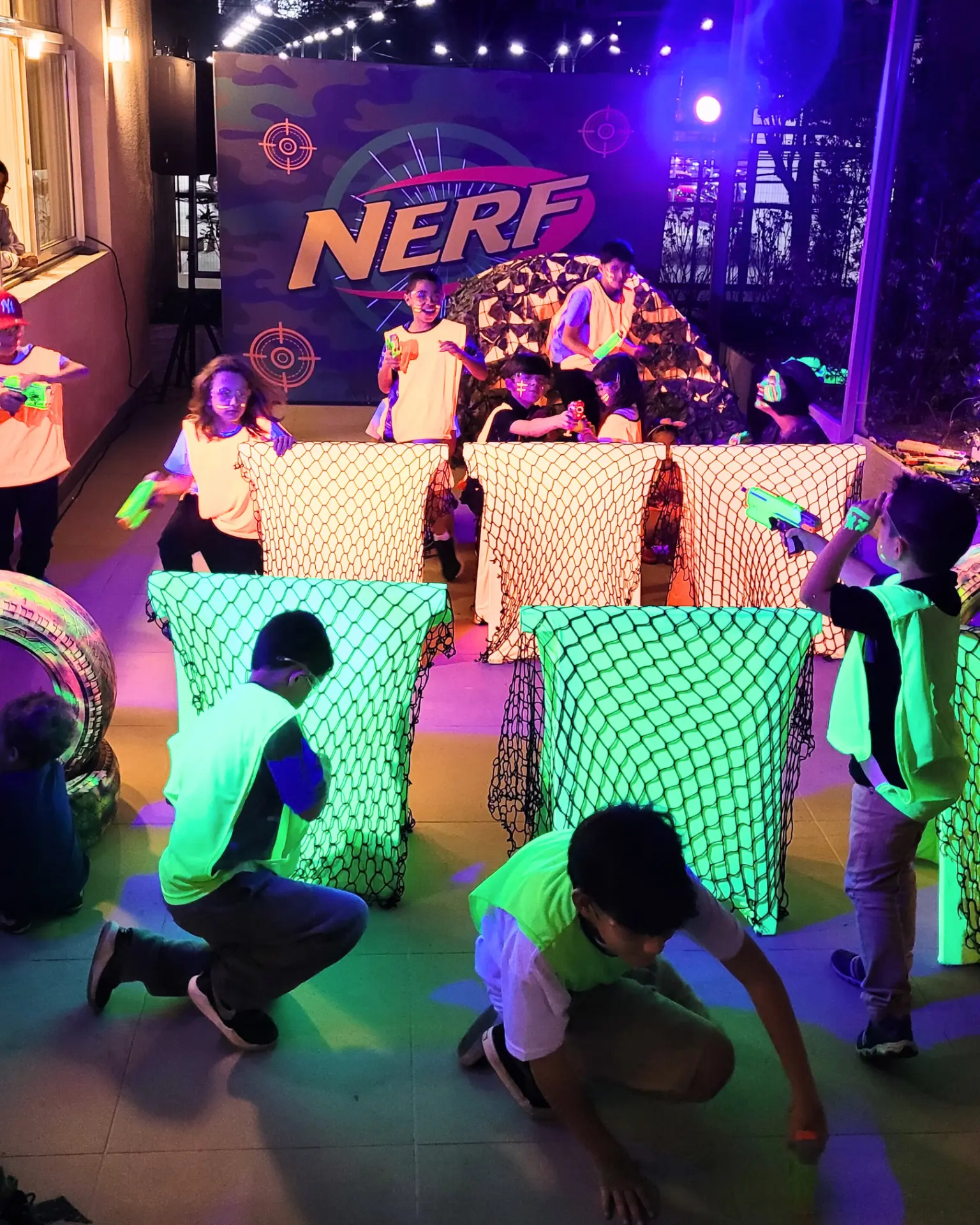 Brincadeiras com Nerf em festa de aniversário à noite, com barreiras e painel iluminados em neon